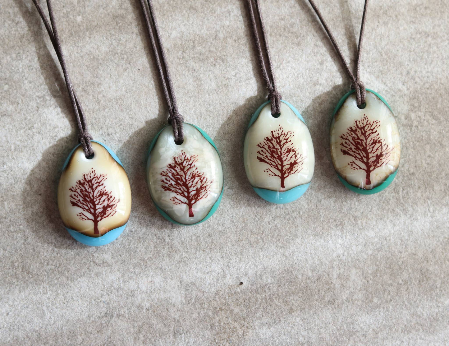 Four necklaces with tree designs on oval pendants against a textured beige background
