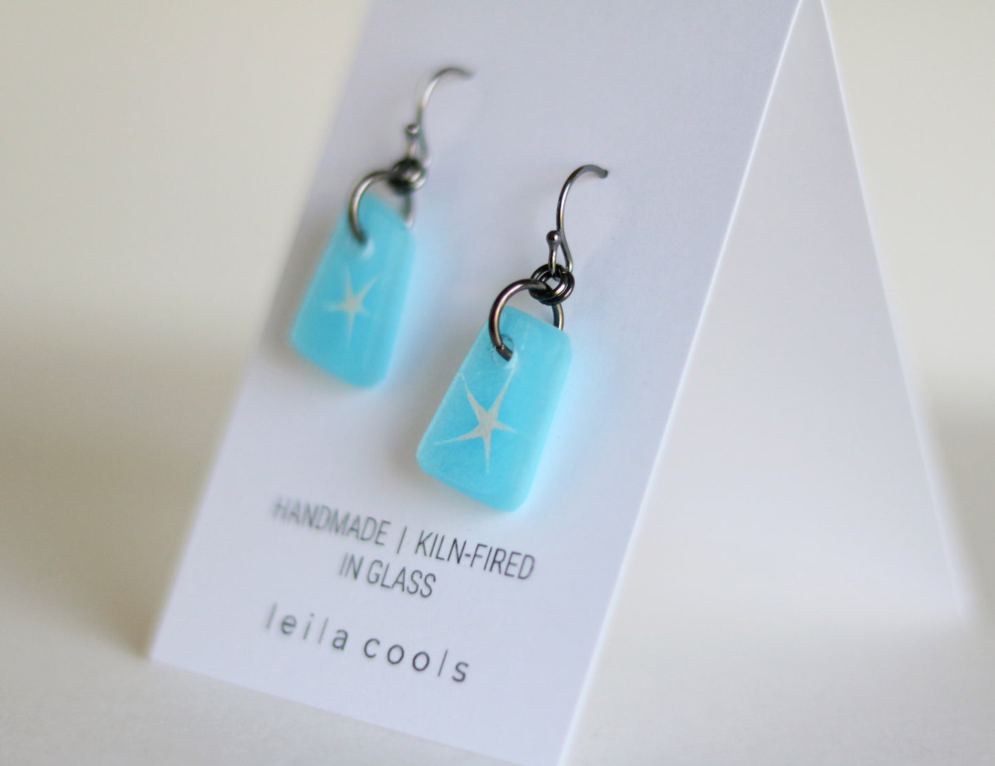 Aqua blue glass earrings with star design on a white card with 'leila cools' branding.
