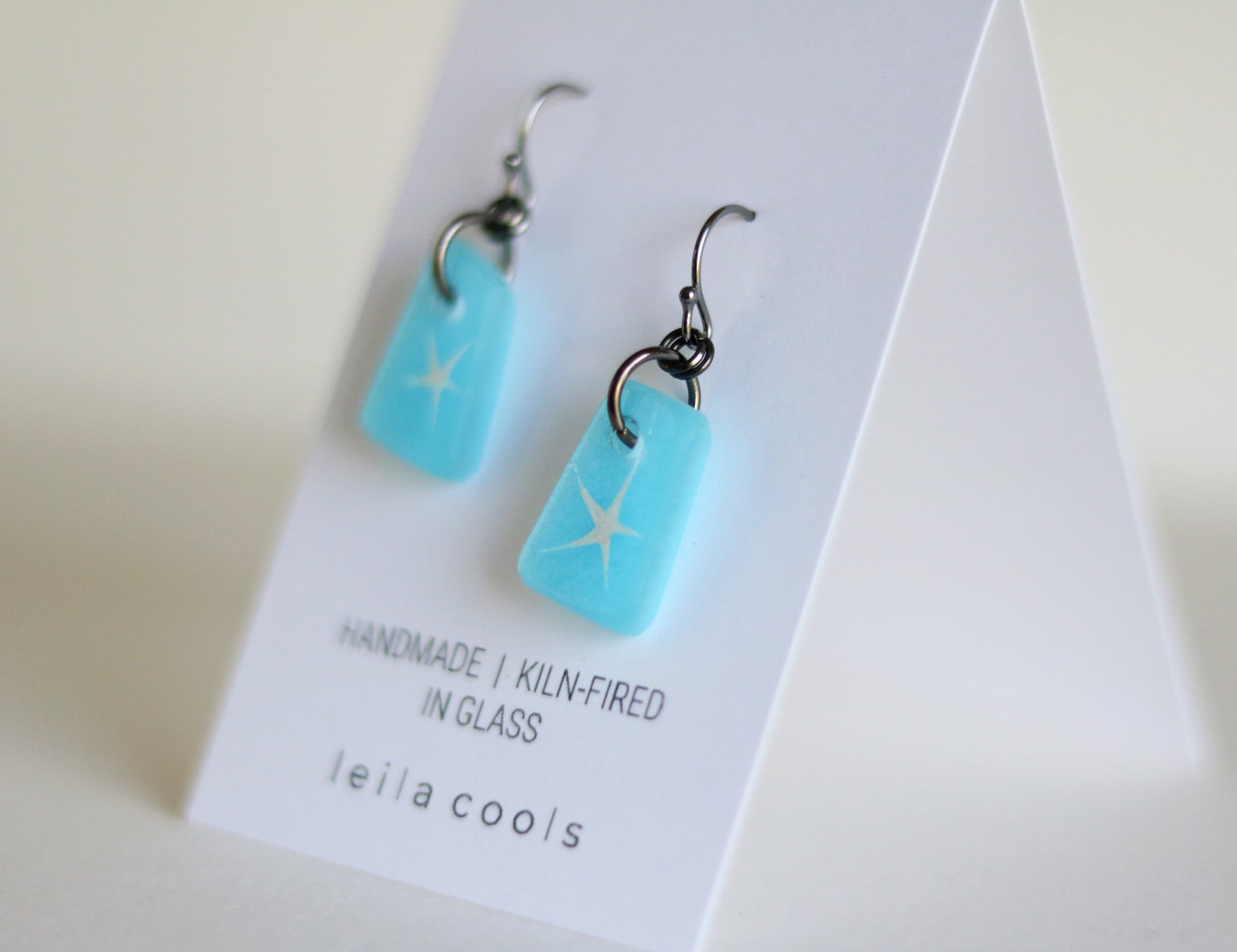 Aqua blue glass earrings with star design on a white card with 'leila cools' branding.