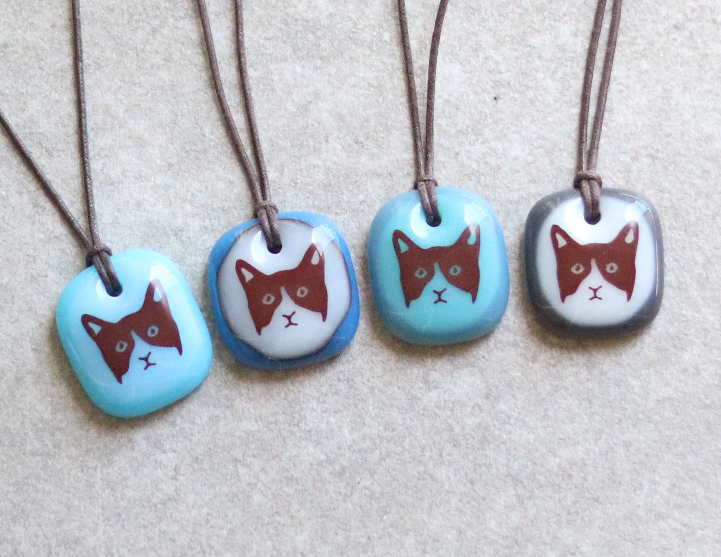 Four cat-themed necklaces with blue and gray pendants on a neutral background