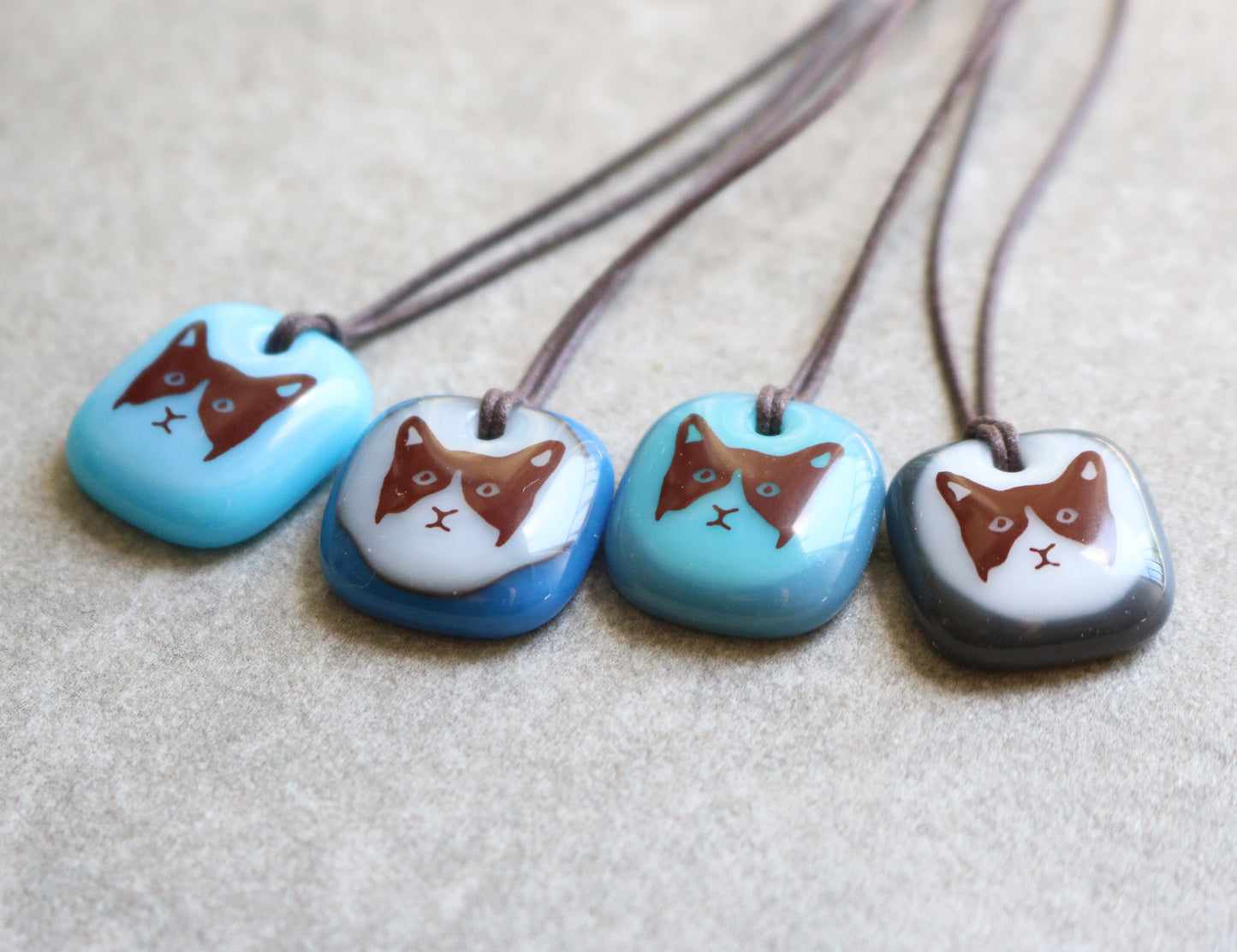 Four glass pendant necklaces with cat designs on a grey ceramic surface