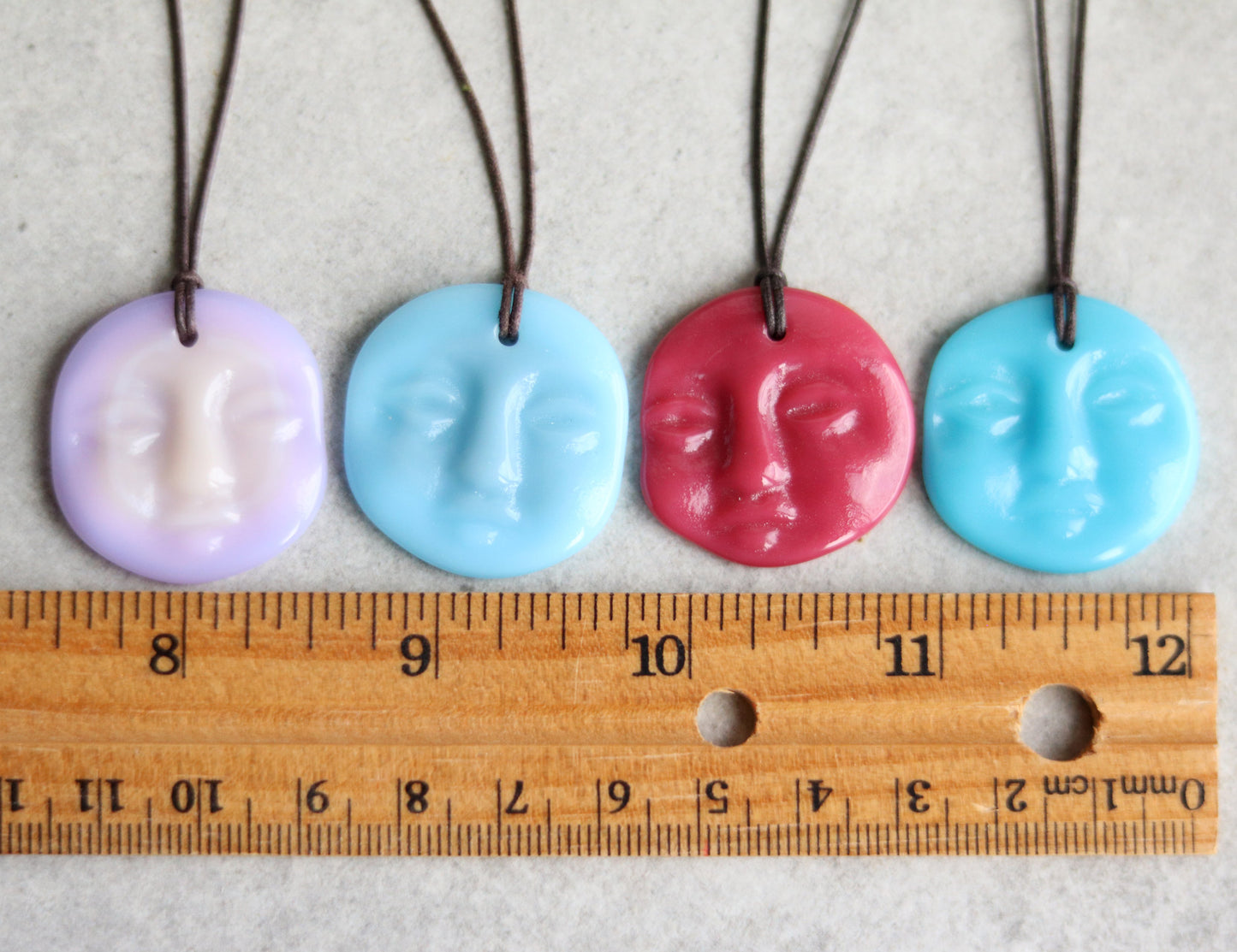 Four colorful face-shaped pendants on strings with a ruler for scale.