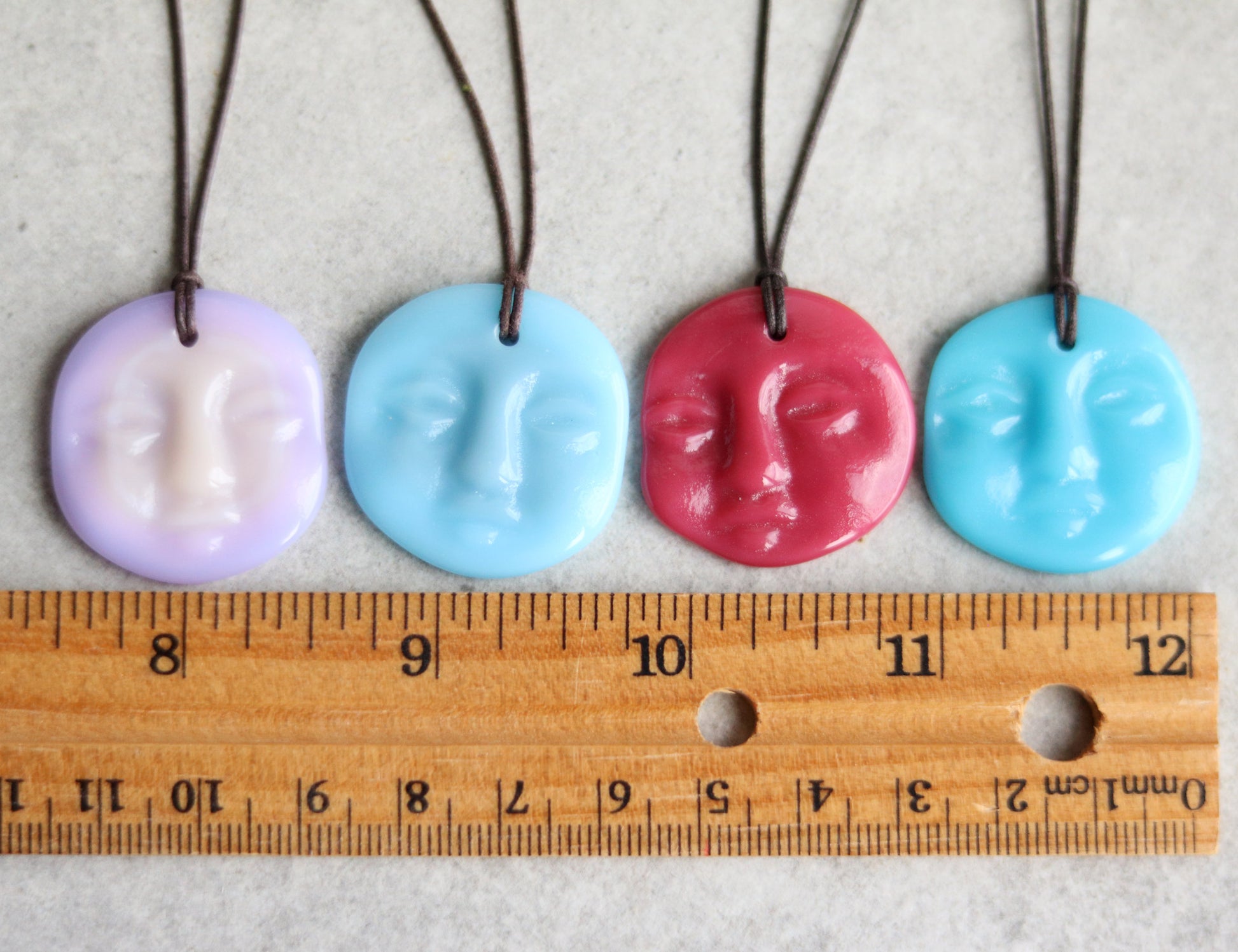Four colorful face-shaped pendants on strings with a ruler for scale.