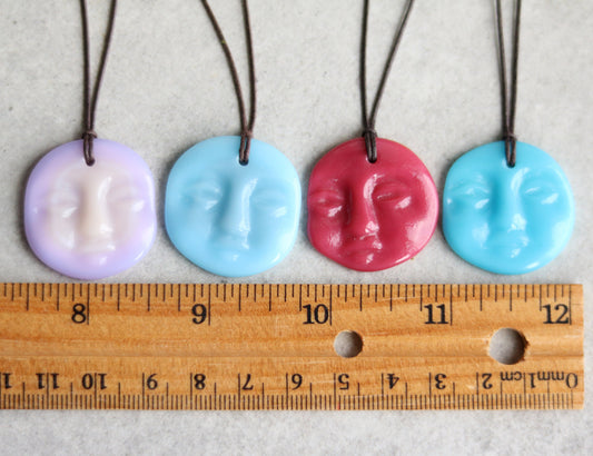 Four colorful face-shaped pendants on strings with a ruler for scale.