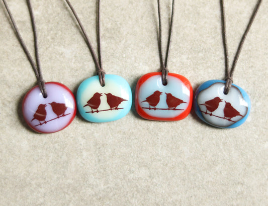 Four necklaces with round pendants featuring bird illustrations on a beige background