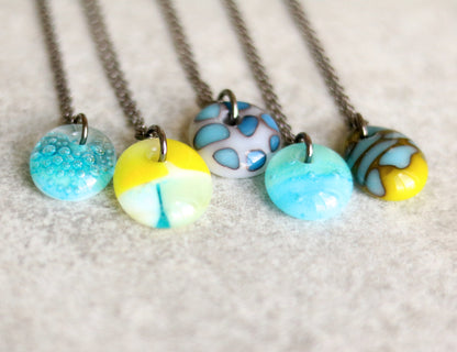 Five chain necklaces with colorful glass pendants on a textured ceramic surface