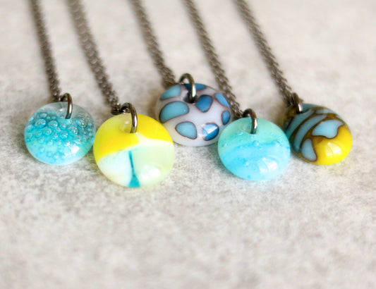 Five chain necklaces with colorful glass pendants on a textured ceramic surface